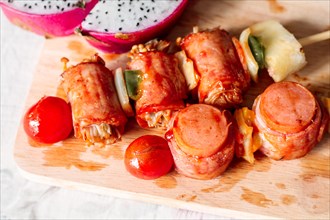 Skewers with bacon-wrapped sausages and vegetables on a wooden cutting board with dragon fruit