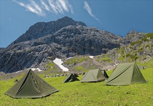 Tents of mountain hunters on a green meadow in front of an imposing mountain backdrop under a clear blue sky