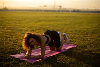 Young curly athletic girl in sportswear performs an exercise on a yoga mat outdoors on the grass during sunset