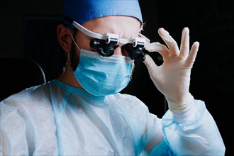 Surgeon wearing binocular magnifying glasses operates on a patient in a dark operating room