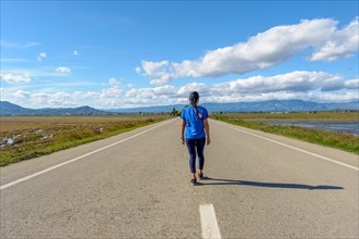 A person is walking down an open road surrounded by nature under a cloud-streaked blue sky