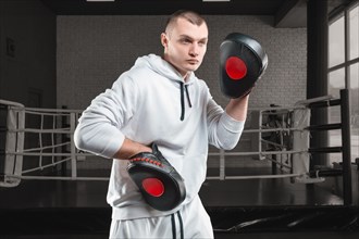 Male trainer in the gym against the background of the ring holds boxing paws. Mixed martial arts concept. High image quality