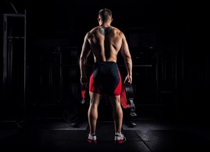 The athlete goes to the gym for training. In his hands he holds sneakers and a belt. Back view