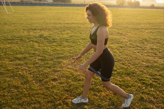 Young curly athletic girl in sportswear performs lunges with resistance band outdoors on the grass during sunset