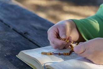 Of a woman's hands praying the rosary beads over a bible in the sunlit countryside