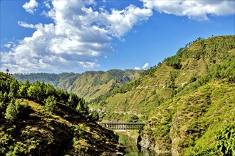 Scenic view of a bridge over Sirot river in a mountainous landscape with lush greenery under a bright blue sky in Manrasa village in the Indian state of Uttarakhand