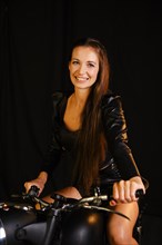 A woman in a black leather jacket and shorts posing on a motorcycle. The image is taken indoors against a dark background