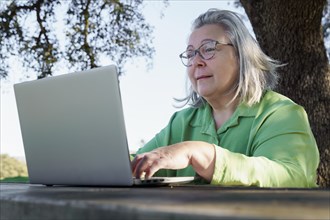 Mature woman with white hair and glasses smiling as she works at her computer on a wooden table in the countryside