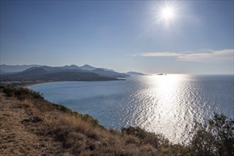 A bright sun over a calm sea with a visible coastline and mountains in the background