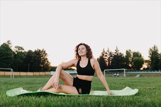 Young curly athletic girl in sportswear resting between exercises while sitting on a yoga mat outdoors on the grass during dawn