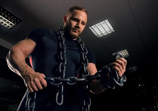 Professional powerlifter stands in the gym with iron chains wound around his arms