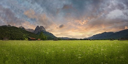 Panoramic view of a green meadow in front of imposing mountains under a cloudy evening sky