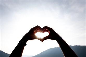 Woman Hands Making a Heart Shape Against the Sky with Sun and Sunbeam and Mountain in Switzerland