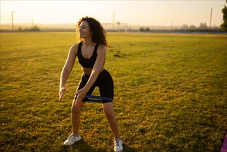 Young curly athletic girl in sportswear performs squats with resistance band outdoors on the grass during sunset