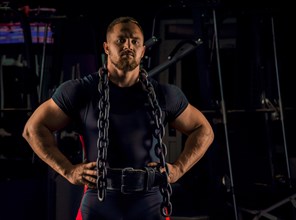 Handsome athlete with a chain around his neck stands in the gym and looks straight ahead
