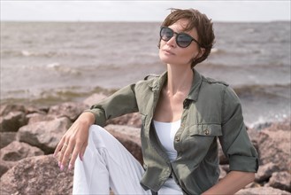 Portrait of a stylish woman sitting on stones in the bay. Windy weather. Happiness