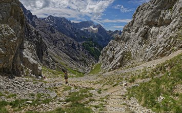 A mountaineer walks down a stony path in a mountainous valley under a cloudy sky