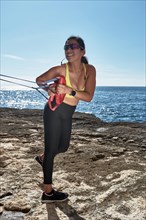 A woman in sporty attire exercises with a resistance band by the sunny seaside