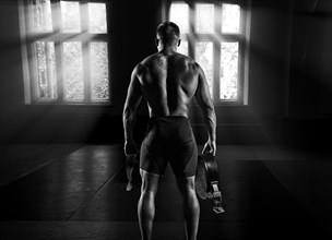 The athlete goes to the gym for training. In his hands he holds sneakers and a belt. Back view