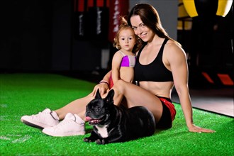 Charming sports mom trains in the gym with her little daughter and a French bulldog.