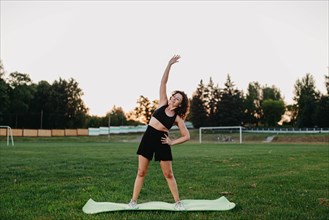 Young curly athletic girl in sportswear performs side bends on a yoga mat outdoors on the grass during dawn