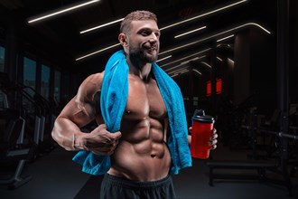 Portrait of a muscular guy with a naked torso and abs. Gym. Blue towel and red shaker. Fitness concept
