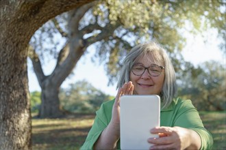 Mature white-haired woman with glasses on a video call with her digital tablet waving in the countryside on a sunny day