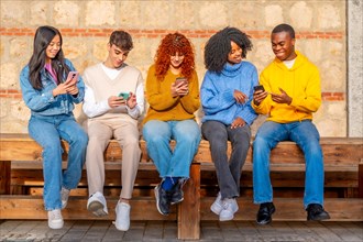 Frontal view of a group of multi-ethnic young friends using phone sitting in a bench in the city