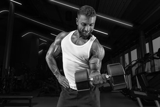 Muscular man in a white t-shirt works out in the gym with dumbbells. Biceps pumping. Fitness and bodybuilding concept.
