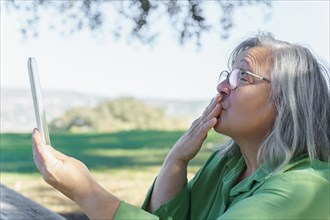 Mature white-haired woman with glasses blowing a kiss to her digital tablet in a video call in the countryside