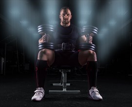 Professional weightlifter sits on a bench in the gym with two dumbbells on his lap