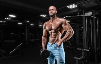 Pumped-up man posing in the gym in jeans with a dumbbell in his hand. Sports