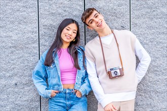 Frontal portrait of two multi-ethnic young friends leaning on a wall carrying a digital camera