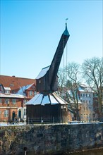 Old crane on the snowy riverbank of the Ilmenau in front of historic buildings under a clear blue sky