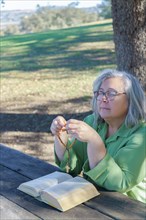 Older white-haired woman with a rosary beads in her hand and a bible praying with her eyes closed sitting at a wooden table in the forest