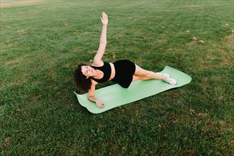 Young curly athletic girl in sportswear performs a side plank exercise on a yoga mat outdoors on the grass during dawn
