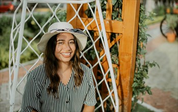 Portrait of a smiling girl in a hat sitting on a white swing in a garden. Lifestyle of girl in hat sitting on a swing looking at camera