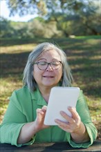 Mature white-haired woman with glasses on a video call with her digital tablet in the countryside on a sunny day