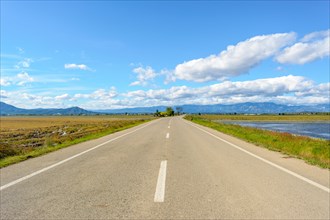 A straight road leading towards mountains under a bright blue sky with clouds