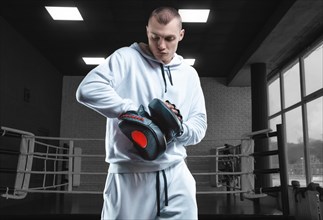 Male trainer in the gym against the background of the ring holds boxing paws. Mixed martial arts concept. High image quality
