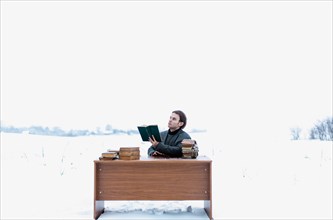 A young handsome intelligent man in a jacket sits at a table in a field and reads ancient books in winter
