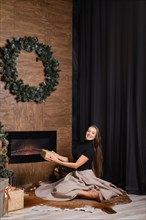Happy woman holds a gift box sitting on the floor by cozy fireplace