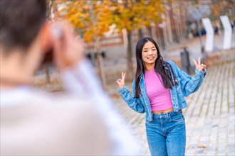 Chinese young woman gesturing victory while a friend taking a photo in the street using a camera
