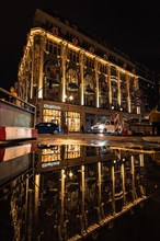 A decorated commercial building at night with Christmas lights reflected in a puddle