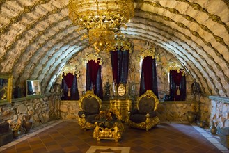 An opulently decorated room with throne chairs under a vaulted stone ceiling and a large chandelier