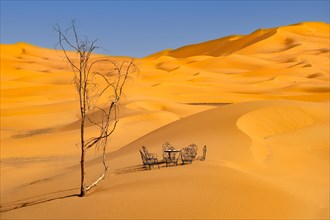 Dead tree and cast iron garden table and lawn chairs in sand dunes of Erg Chebbi