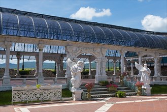 A pergola with outdoor sculptures against a mountainous landscape and cloudy sky