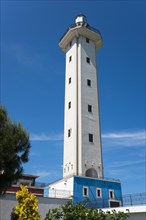 A white lighthouse with a blue base against a blue sky surrounded by green and yellow