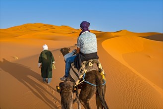 Cameleer and Western tourist riding dromedary camel in sand dune of Erg Chebbi
