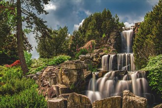 A picturesque waterfall flows over rocky steps surrounded by lush vegetation and dramatic skies,